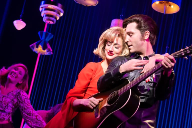 man with guitar standing next to a woman in a red dress with a blue background
